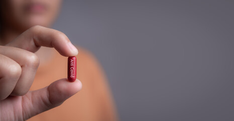 Female hand holding a medical red capsule pill between fingers against gray...