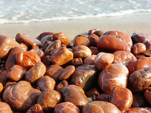 Unique red jasper beach. Many pieces of red jasper polished by the sea. Wet sea stones on a blurred background of the oncoming wave.