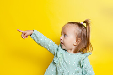 Portrait of a cute baby girl with blue eyes on a yellow background points to the side. A place for text.