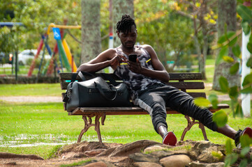 African black man in sunny afternoon, sitting on bench, using smartphone, with his black bag beside him, relaxing outdoors in park, healthy life.