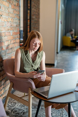 Young female scrolling in smartphone while sitting in front of laptop in cafe or office