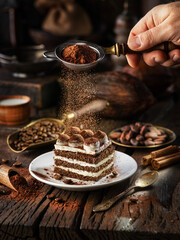 Dusting tiramisu-like cake with cocoa powder. Still life with slice of cake and coffee and cacao...