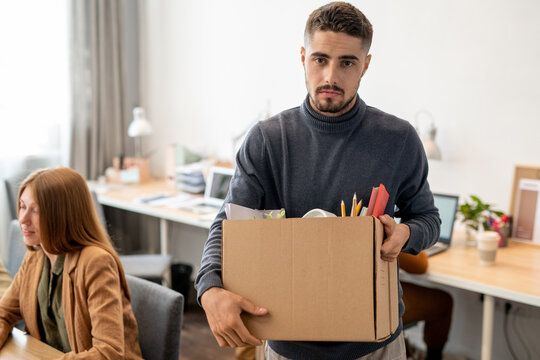 Young unhappy employee holding box with office supplies while standing in front of camera