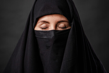 Naklejka premium Close up portrait of young, adult woman in black burqa with hidden face, isolated on black background.