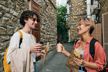 Happy young dates with drinks and snacks looking at one another while standing in the street