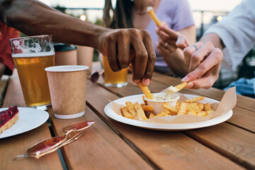 Hands of multiracial friends putting french fries into spicy sauce while eating it in outdoor cafe