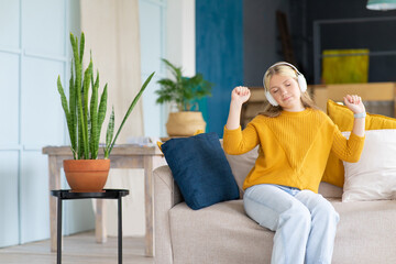 Teenage girl in yellow sweater uses wireless white headphones for listening to music home. Home interior background with copy space. Home relaxation.