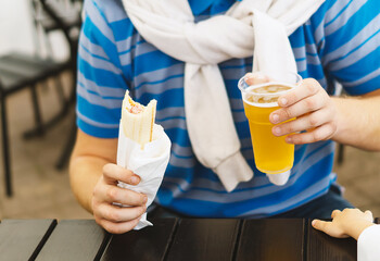 Men's hands hold a hot dog and a glass of golden beer. A young man is sitting at a table in an outdoor cafe, drinking light beer and eating a hot dog. The guy enjoys food and drinks. Harmful lifestyle