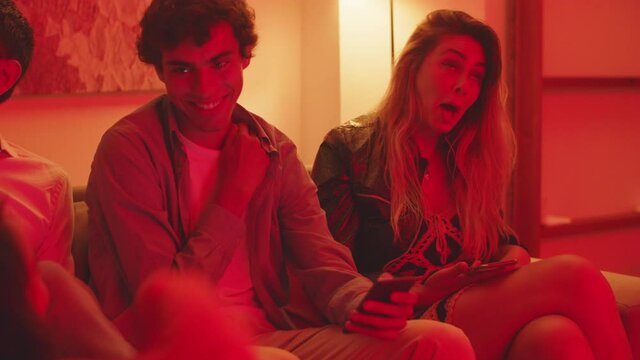 Medium shot of girl sitting on couch with friends and yawning. Handheld shot of group of friends having night party in apartment, laughing, chatting amd spending good time. Home party concept