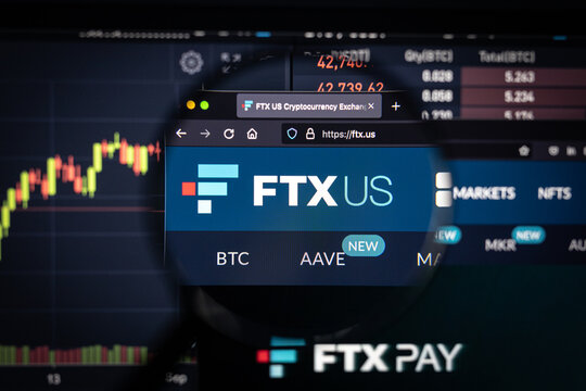 FTX US cryptocurrency exchange logo on a website with blurry stock market developments in the background, seen on a computer screen through a magnifying glass