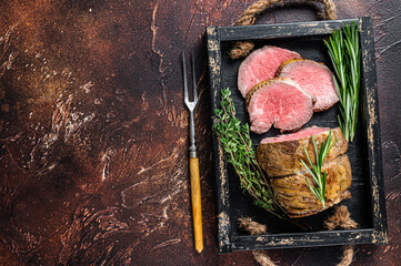 Roast beef round fillet meat in a wooden tray with herbs. Dark background. Top view. Copy space