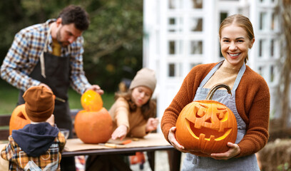 Smiling woman with jack-o-lantern smiling at camera while carving pumpkins with family in backyard
