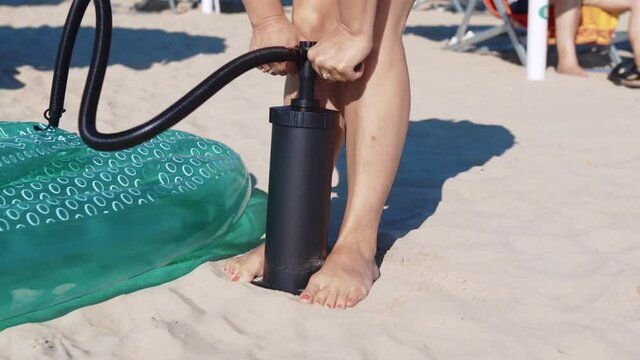 Woman inflating green air mattress with black manual pump at the sea beach, female preparing trendy inflatable float mattress for swimming on water surface. Woman enjoying summer vacation and relax