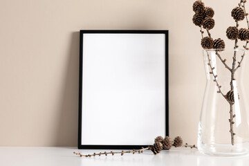 Black photo frame mockup and branch with pine cones on beige wall background. Autumn or winter background. Front view