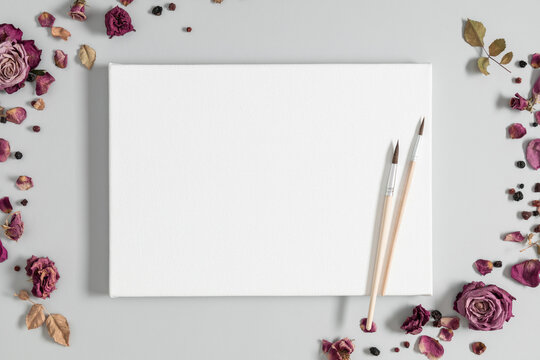 Blank white canvas mockup with paint brushes with dry roses on gray background. Flat lay, top view, copy space