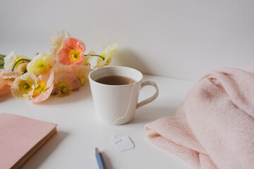 Close up of white cup with tea bag on table with flowers, journal, pen and shawl - self care concept (selective focus)