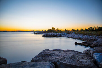 The rocky shoreline and calm, glassy water of Lake Ontario are seen during a colourful sunset in...