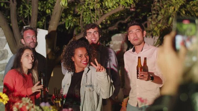 Back view of woman taking photo of friends at outdoor party. Handheld shot of group of multicultural people posing for camera, drinking beer, smiling and having fun. Night party, friendship concept