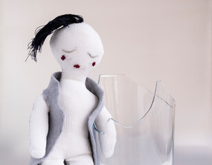 Muñeco de trapo con expresión triste, fondo blanco y vaso de vidrio roto 