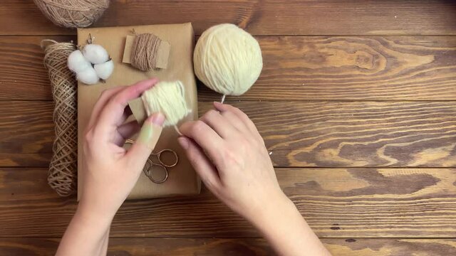 Woman wind white yarn on piece of cardboard. Box in craft wrapper, twine, cotton and scissors on wooden table. Handmade crafting decorations of gift for Christmas or Birthday. New Year mood.