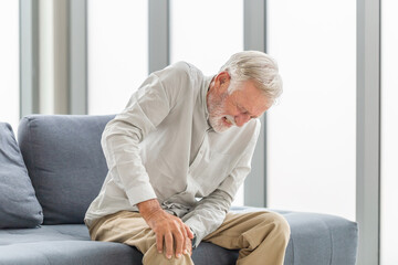 Old man suffering from knee pain sitting sofa in the living room, Elderly man suffering from knee pain while sitting on the sofa - obrazy, fototapety, plakaty
