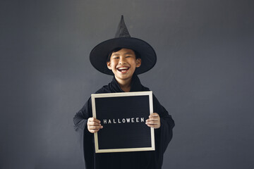 Cute Asian boy celebrating Halloween by wearing witch costume and holding Halloween decoration accessories