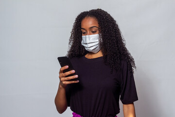 Mulher jovem negra com cabelos cacheados usando máscara contra covid e usando aparelho celular