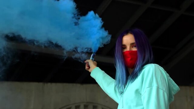 attractive blue-haired woman in a mask walking with blue smoke granade in her hand and looking to the camera, women's rights concept