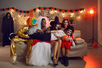 Obraz na płótnie Canvas Group of multiracial adult friends having fun at Halloween costume party in decorated living room. Young people disguised as spooky characters laughing sitting on sofa with one man lying on their laps