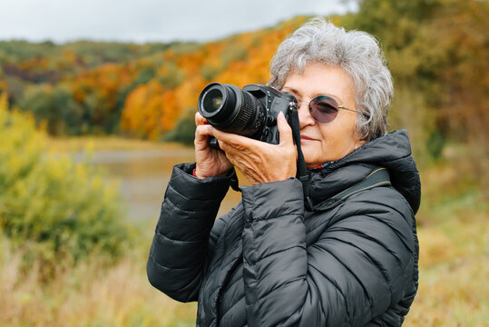 Senior woman with glasses using digital camera, retired woman photographing autumn landscape in front of forest and lake outdoors. Elderly woman photographer tourist, retired hobby