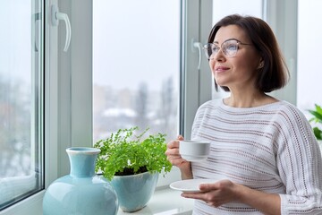 Middle aged beautiful woman with glasses with cup of coffee at home in winter autumn season