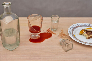 A broken glass, red tomato juice spilled on the table next to a bottle of vodka