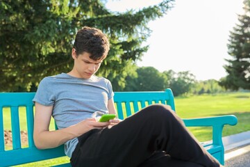 Serious guy teenager using smartphone, having rest, sitting on bench in park