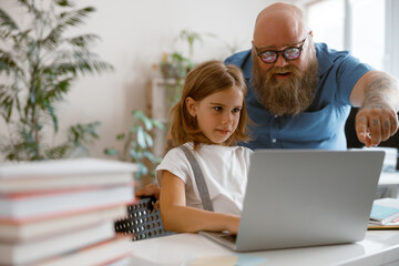 Bearded man helps little girl to work on laptop during lesson in light room