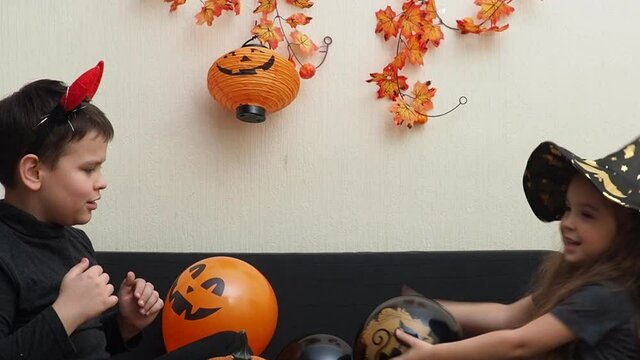 boy and girl, brother and sister play with balloons in fancy dress at home preparing for halloween. Girl in a witch hat. inflate balloons, prepare decorations. girl with devil horns carves bats 