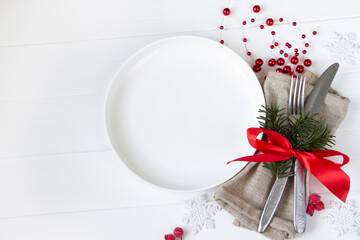 Christmas table setting-white plate, table fork and knife set with napkin, fir branch and red ribbon, white snowflakes, red berries on white wooden board