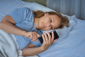 Senior woman using mobile phone while lying in bed. Pensioner suffers from insomnia
