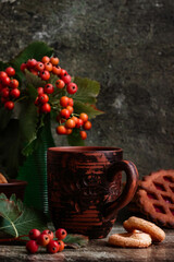 A bouquet of red berries in a green glass vase on a dark wooden background, next to it is a brown ceramic mug with hot tea and round cookies and red berries. Autumn homely cozy composition for tea dri