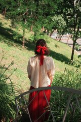 Young girl in a romantic outfit with her back to the camera in the park. Back view. Red bow in...