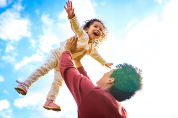 happy african american little fearless girl flying outdoor on father's hands in summer park