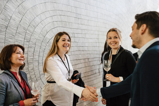 Happy pregnant businesswoman greeting businessman while doing handshake standing by colleagues at convention center