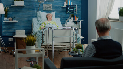 Retired patient with disease sitting in hospital bed with heart rate monitor and IV drip bag in nursing home. Ill woman waiting for healing treatment while senior man assisting sick wife