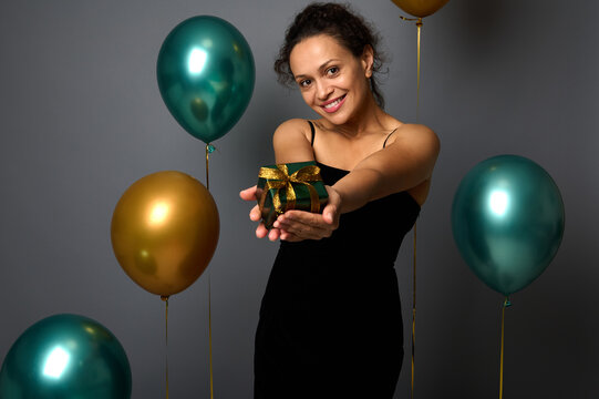 Mixed race cute woman in black dress holds a Christmas gift in outstretched hands and shows it, looking at camera, isolated over gray background with gold and green air balloons, copy space for ad