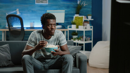 African american man enjoying comedy on television while eating popcorn at home. Black person...