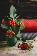 a bouquet of red berries in a glass vase, warm knitted things are lying on a wooden table on a dark...
