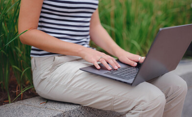 Naklejka premium happy asian female student working with laptop in modern university campus park. woman in striped T-shirt and light trousers works at gray laptop. Hands closeup