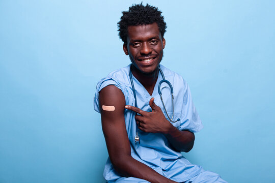 African American Nurse Pointing To Vaccine Shot Bandage While Looking At Camera And Smiling. Black Medical Assistant Showing Adhesive Plaster After Getting Vaccinated Against Coronavirus