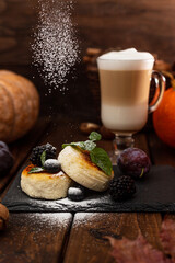 Two cheesecakes and a glass mug with latte stand on a wooden table on a black stand. Powdered sugar is poured on top of the cheesecakes. Decorated with berries and mint