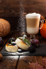 Two cheesecakes and a glass mug with latte stand on a wooden table on a black stand. Powdered sugar is poured on top of the cheesecakes. Decorated with berries and mint