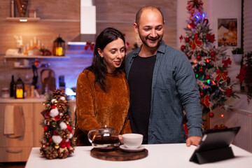 Joyful couple discussing with remote friends during online videocall meeting conference using laptop computer standing at table in xmas decorated kitchen. Haapy family celebrating christmas holiday